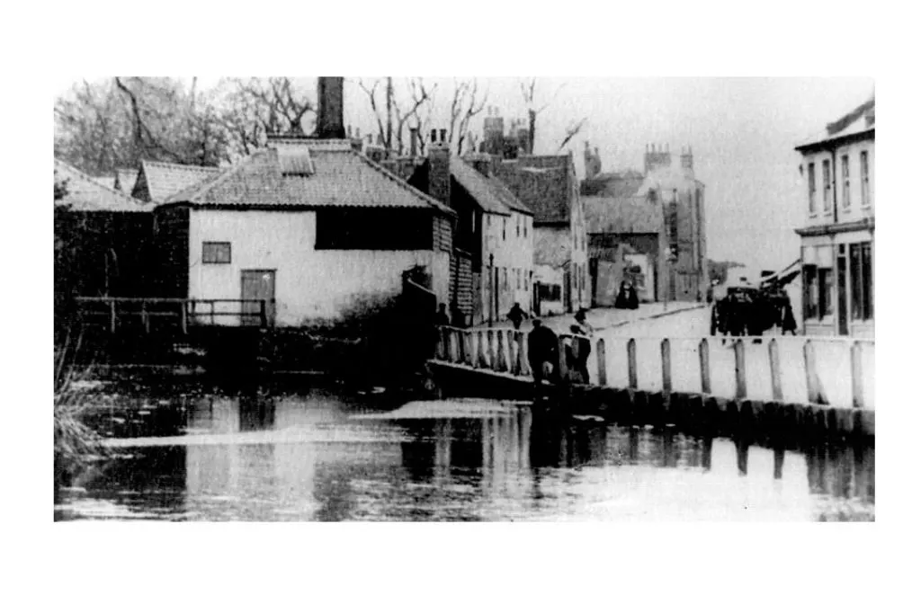 Black and white photo of Merton High Street in 1880, showing small buildings and people near the River Wandle.