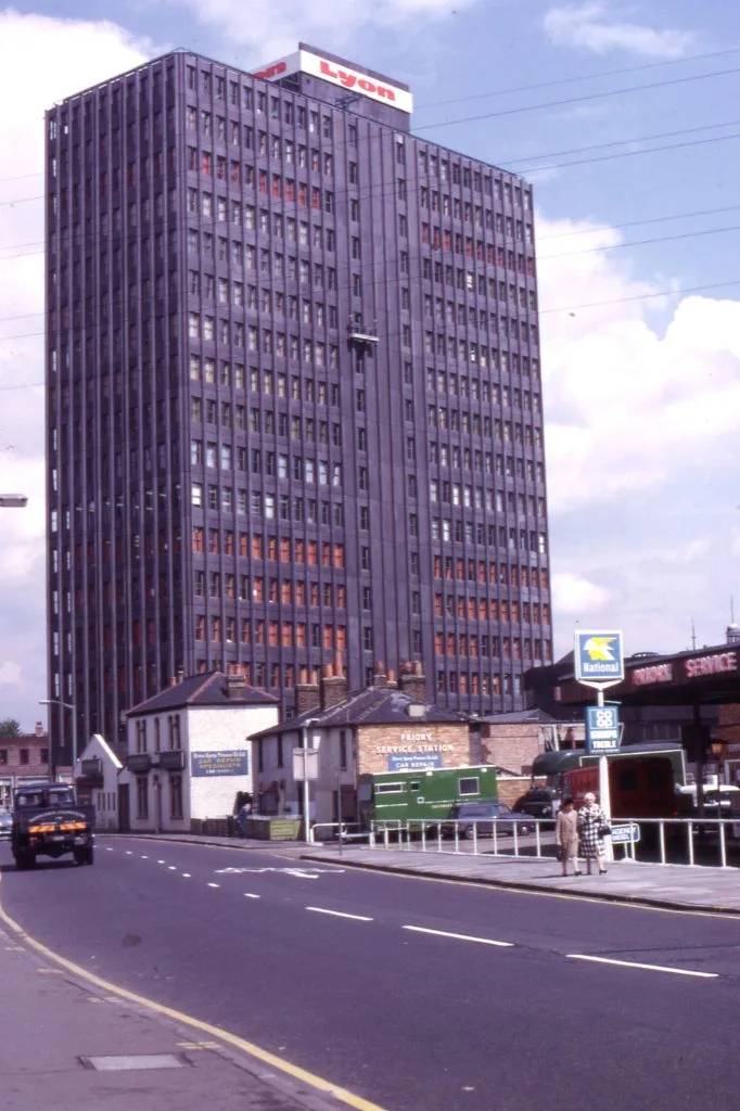 Tall dark tower block in Colliers Wood with the sign "Lyon" at the top, viewed from the street with cars and pedestrians.