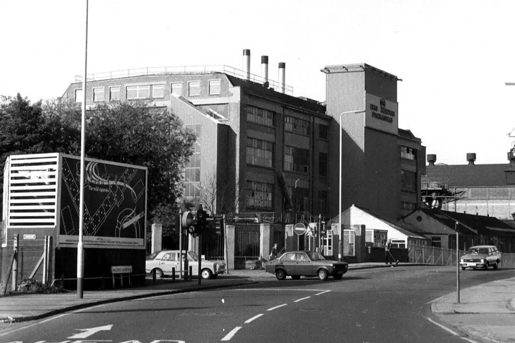 Black and white photo of the large Merton Board Mill factory building with chimneys, cars, and street signs in front.