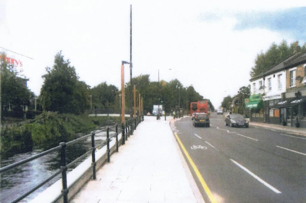 Photo of Merton High Street showing a wide pavement beside the River Wandle, cars on the road, and shops on the right.