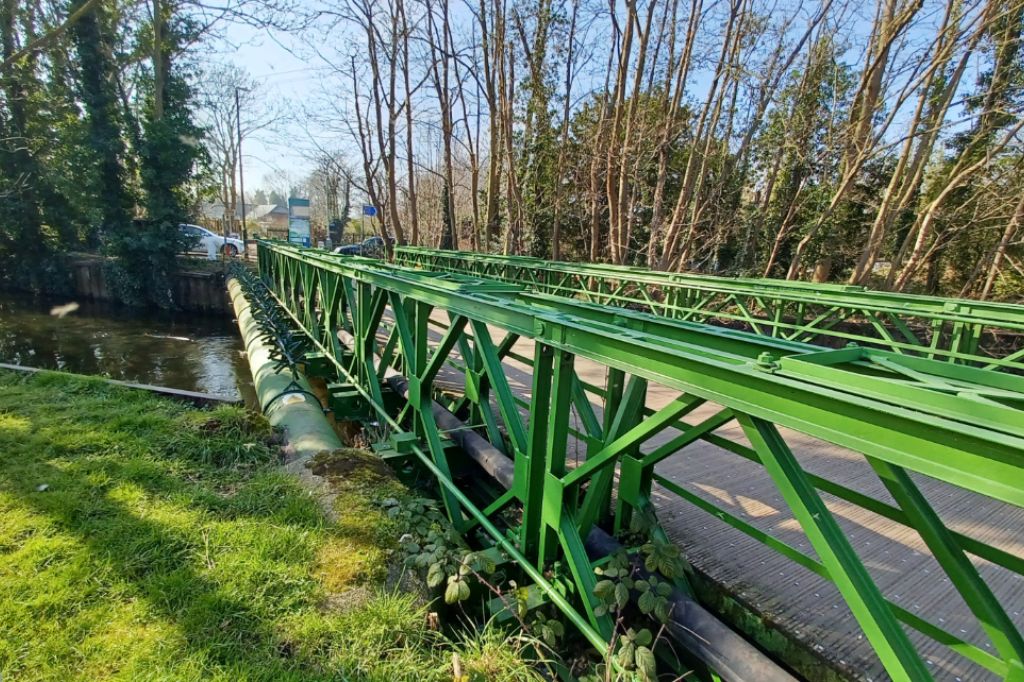 Green metal footbridge crossing the River Wandle with trees and parked cars in the background.