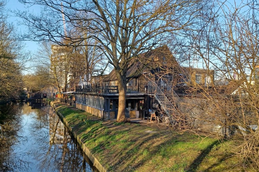 Riverside view of a historic building at Merton Abbey Mills with outdoor seating.