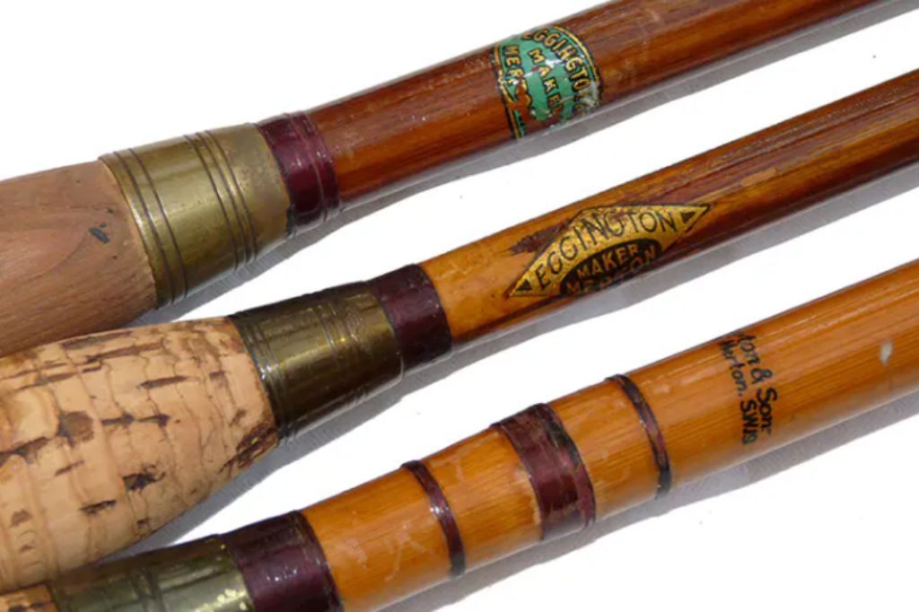 A close-up photograph of three vintage bamboo fishing rods, handcrafted by Eggington & Sons, a fishing rod maker based in Merton. The rods feature brass fittings, varnished cane, and cork or wooden handles. Each rod is labeled with 'Eggington Maker Merton' or 'Eggington & Sons, Merton SW19' in decorative lettering. The rods display fine craftsmanship, with intricate thread bindings and polished metal accents.