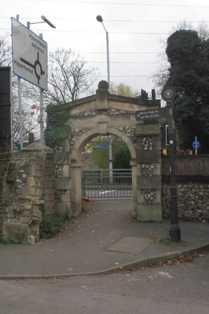 A reconstructed medieval-style gateway at Merton Priory, made of flint and stone, with a pedestrian path leading through it.