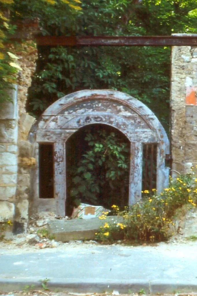 An old photograph of a medieval gateway at Merton Priory, showing a weathered wooden door within a stone arch, partially overgrown with vegetation.