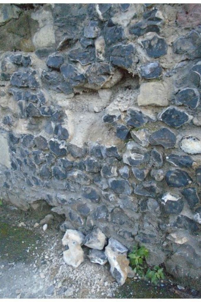 Close-up of Merton Priory’s crumbling flint and stone wall, with missing sections and fallen debris at its base.