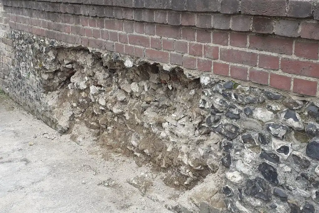 A large hole in Merton Priory’s historic flint and stone wall, with crumbling mortar beneath a layer of Victorian brickwork.